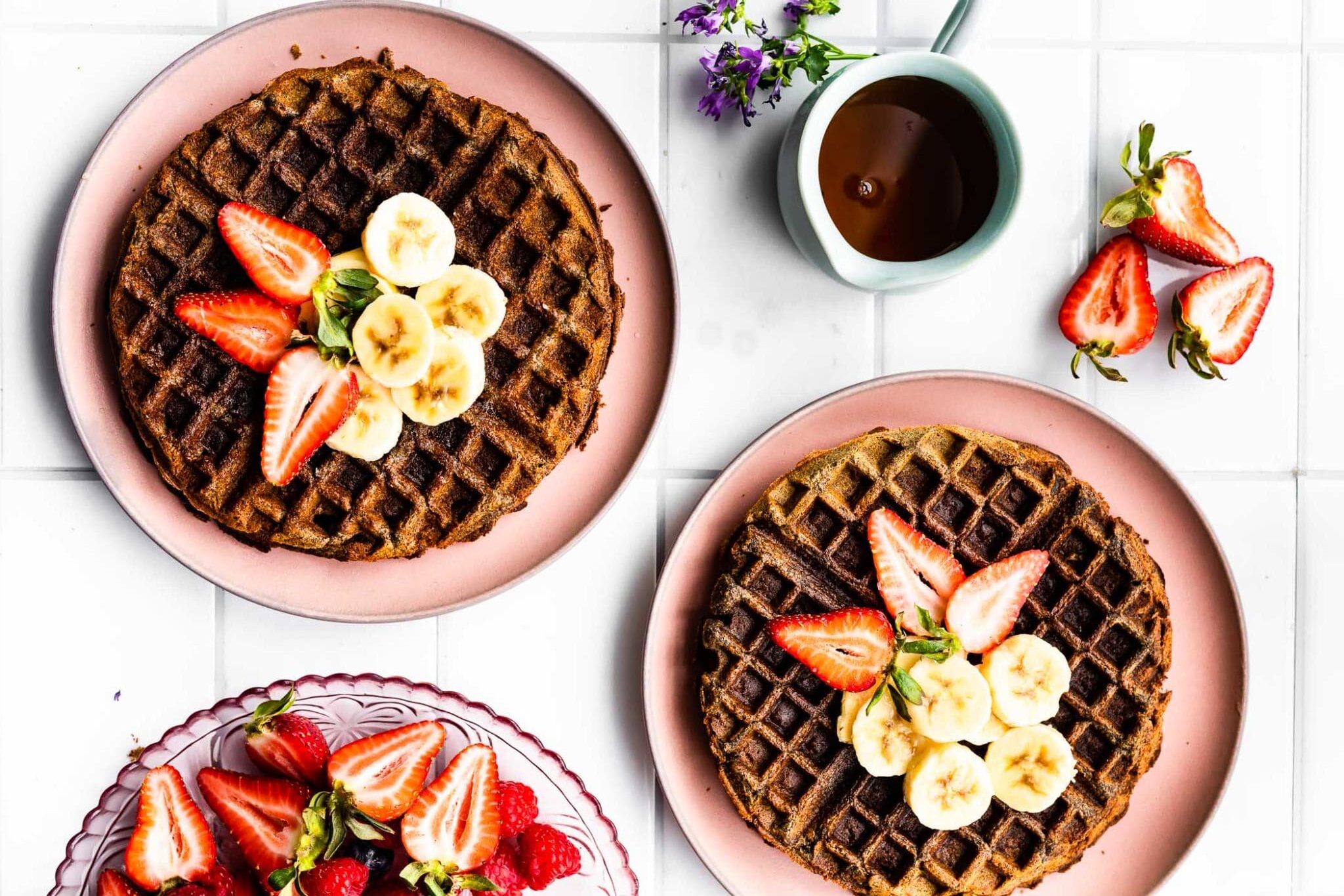 Overhead view two pink plates holding a round vegan buckwheat waffle each, topped with sliced strawberries and bananas.