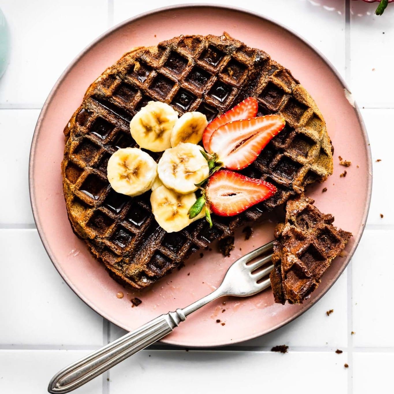An overhead photo of a vegan banana buckwheat waffle, topped with banana and strawberry slices, on a light pink plate.