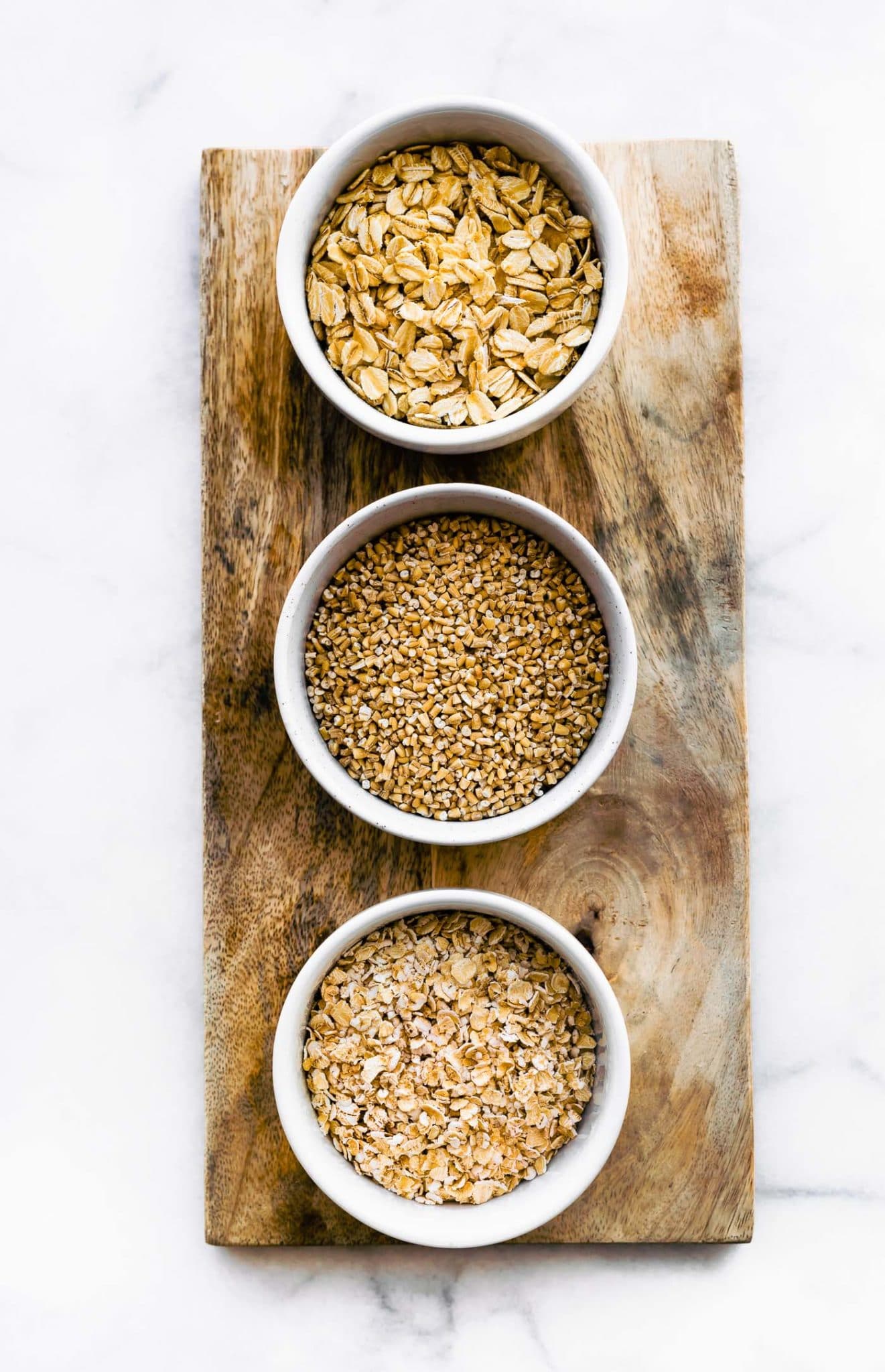 overhead image of three white bowls filled with rolled oats, quick oats, and steel cut oats on a wooden board