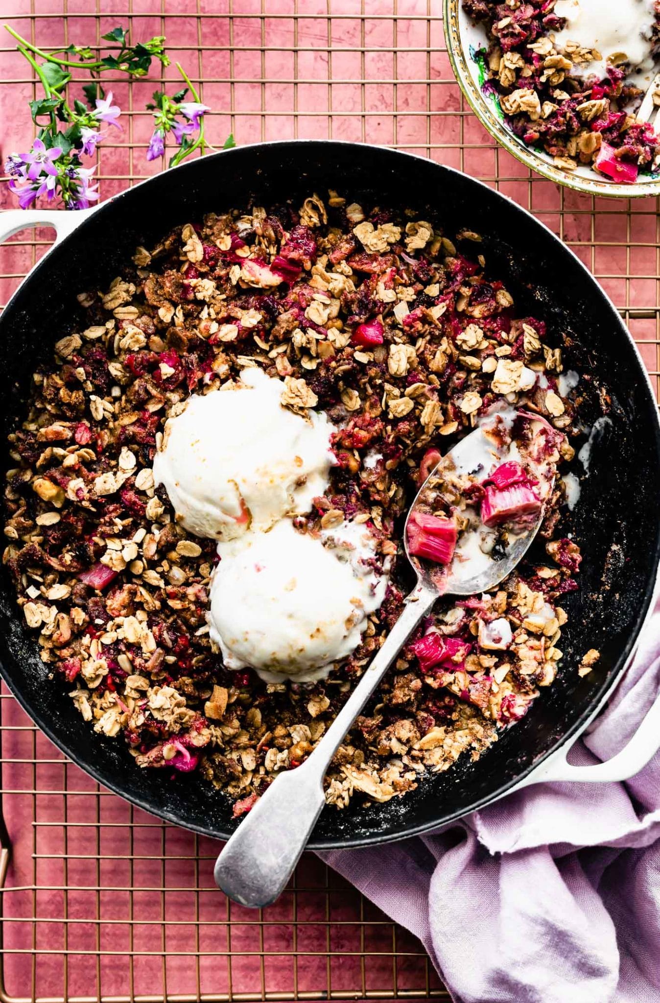 Overhead view berry rhubarb crumble in skillet with two scoops vanilla ice cream melting into top.