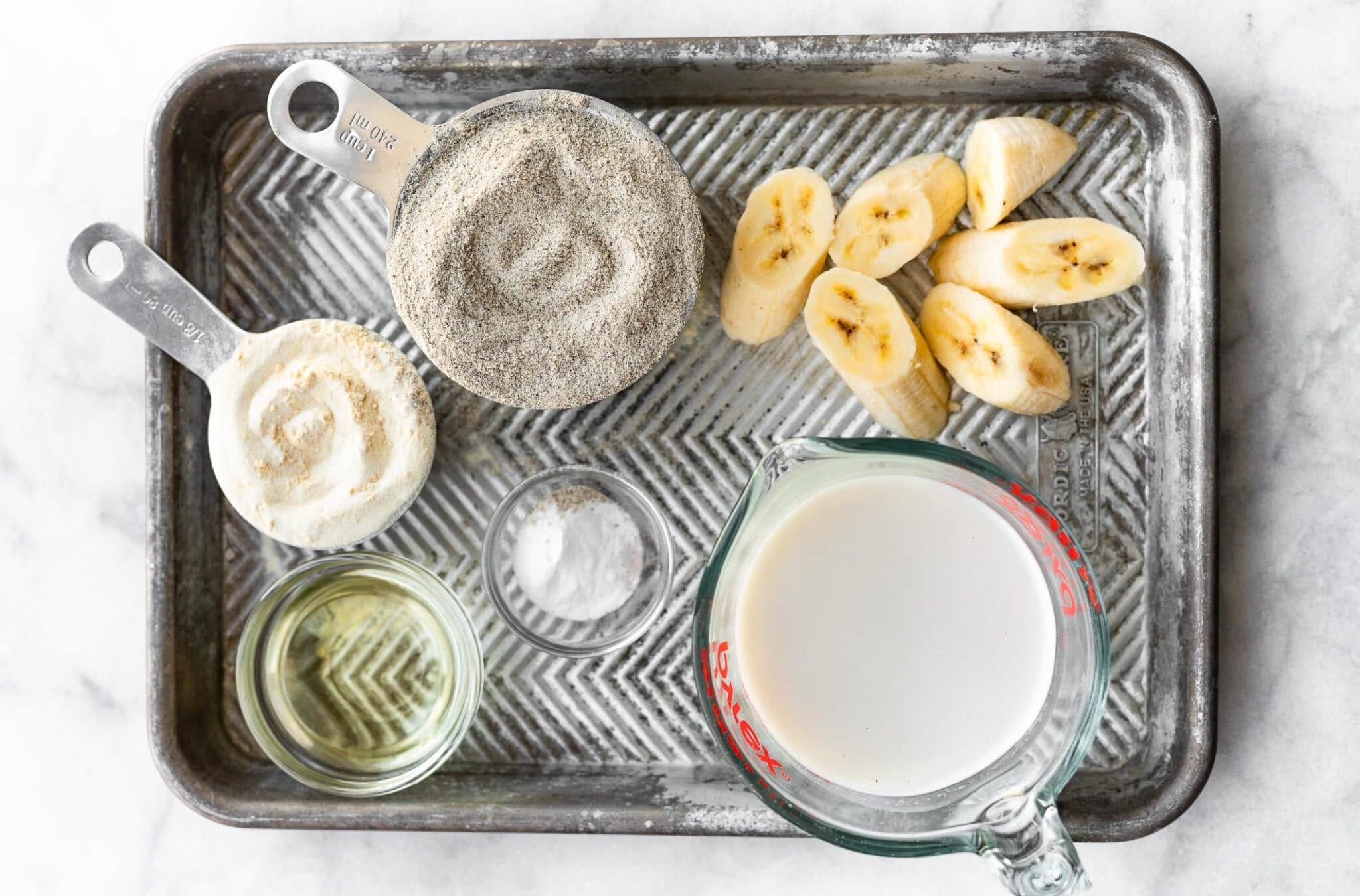 overhead view of non-dairy milk, sliced bananas, buckwheat flour, and other ingredients on a baking sheet to make vegan buckwheat banana waffles