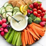 a snack platter with celery sticks, carrot sticks, broccoli florets, strawberries, grape tomatoes, gluten free crackers, lemon wedges, cucumber slices, radishes, and a small bowl of vegan ranch dressing with a spoon in it in the center