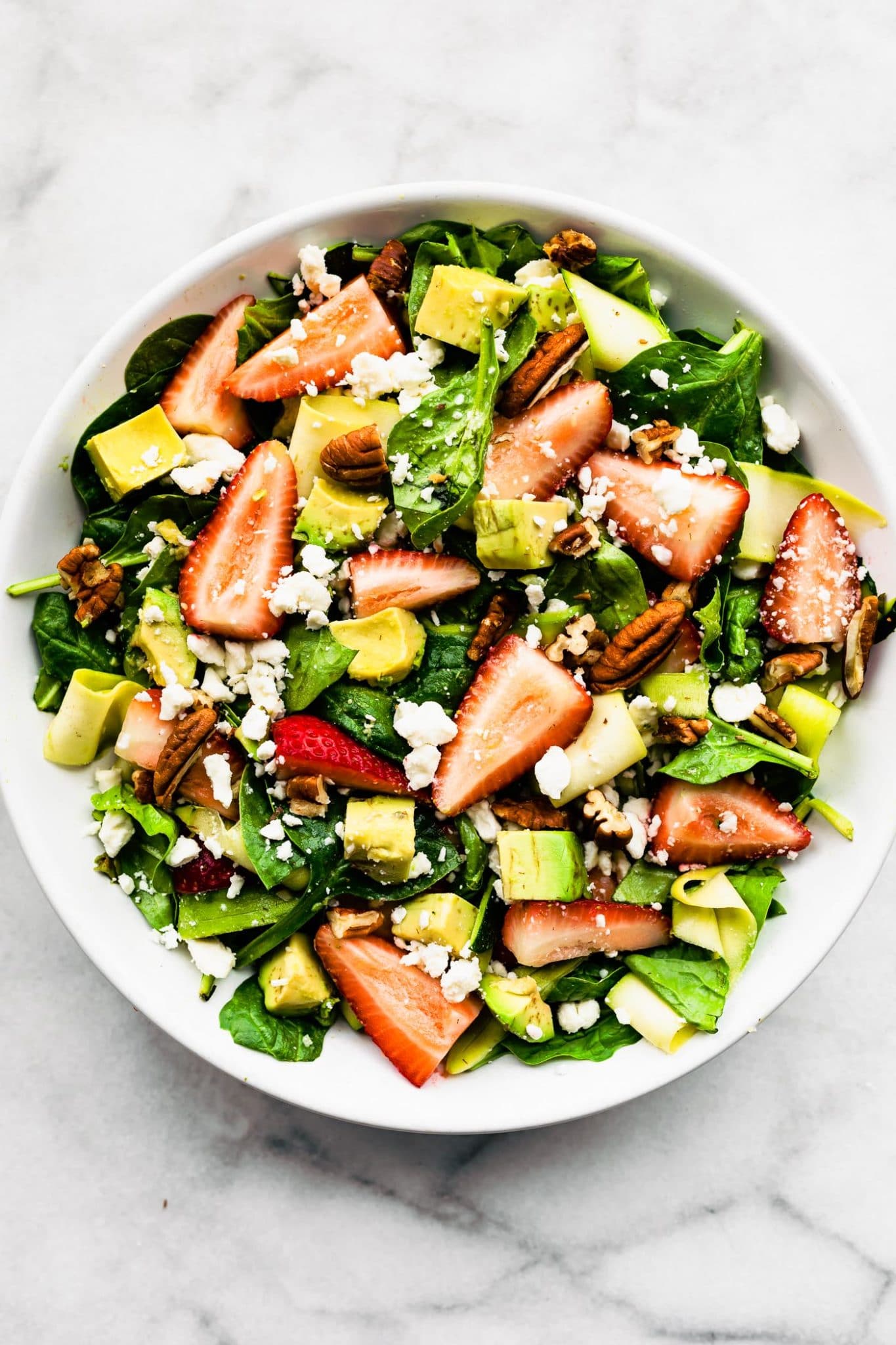 overhead image of a summer strawberry salad recipe including spinach, strawberries, avocado, feta, and pecans in a white bowl