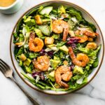 Overhead view spicy shrimp Caesar salad in white and brown bowl topped with croutons and avocado pieces.