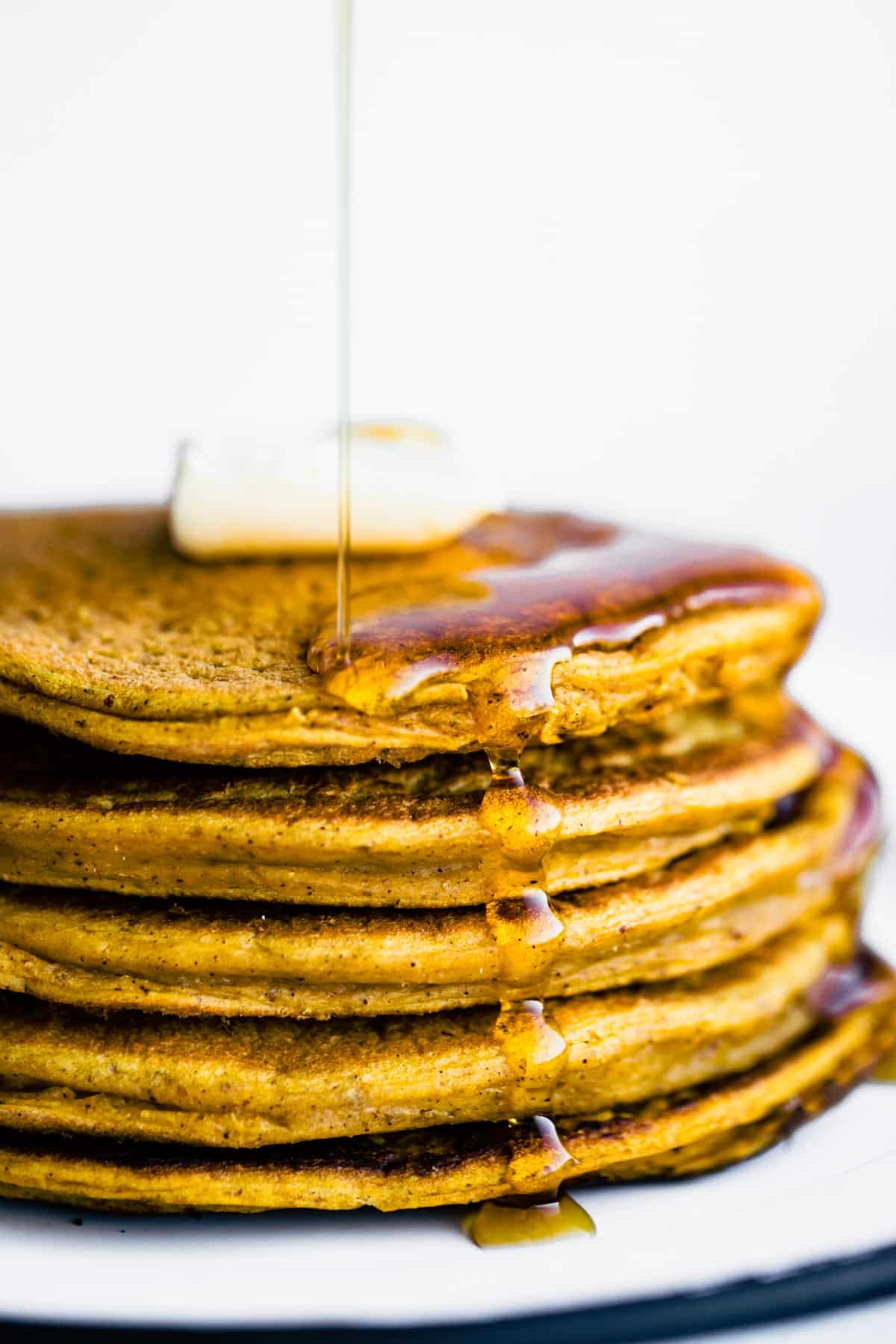 a stack of vegan pumpkin pancakes topped with a pad of butter being drizzled with maple syrup