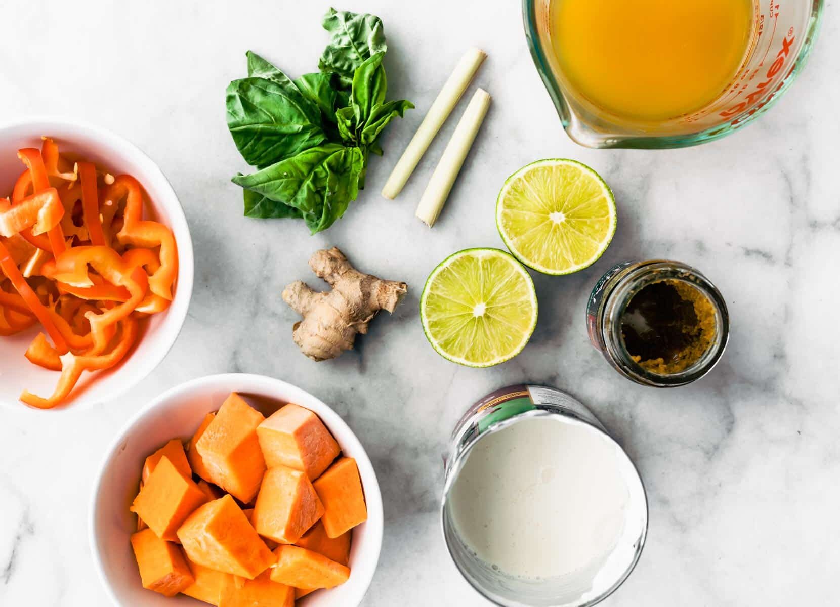 Overhead shot of ingredients on a counter top - lime split in half, ginger root, red peppers cut and in a bowl, sweet potatoes chopped up in a bowl, broth, coconut milk in a can and a few leaves of basil.