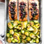 A sheet pan with three salmon filets topped with a blueberry mixture and Brussels sprouts.