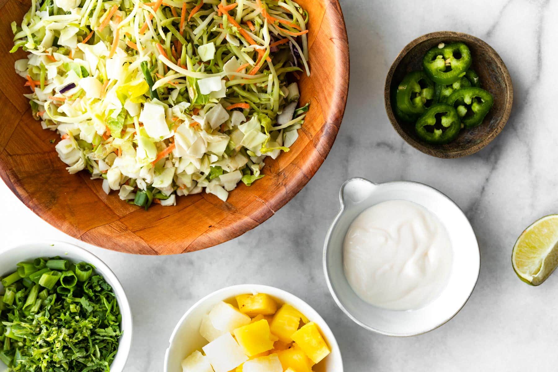 Overhead image of bowls of shredded cabbage and broccoli slaw, scallions, pineapple chunks, mayo, jalapeños, and a lime wedge.