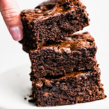 A hand grabbing a gluten free brownie from a stack of three brownies on top of each other on a plate.