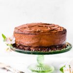 A gluten free chocolate cake iced with chocolate frosting on a cake stand surrounded by chocolate chips.