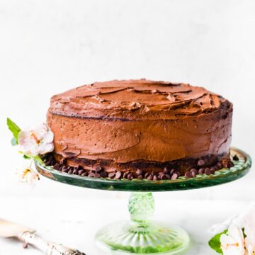 A gluten free chocolate cake iced with chocolate frosting on a cake stand surrounded by chocolate chips.