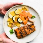 An overhead view of hands holding a white plate with jerk salmon and roasted veggies.