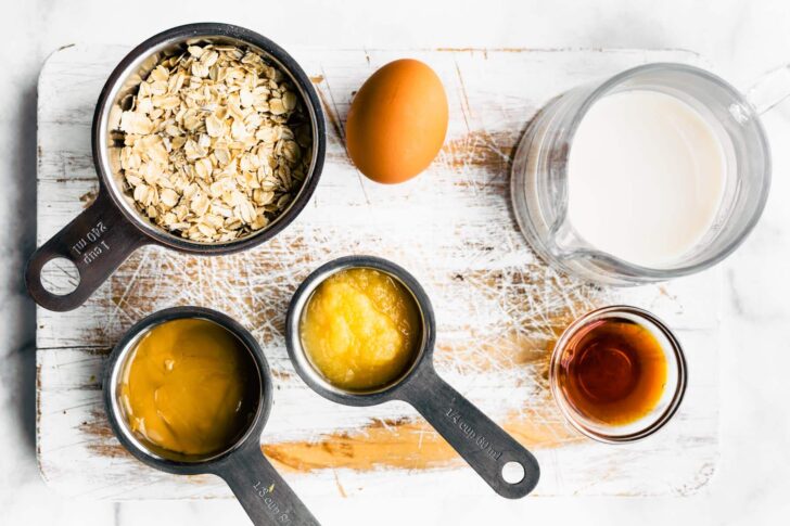 Measuring cups filled with oats, peanut butter, and apple sauce on a cutting board.
