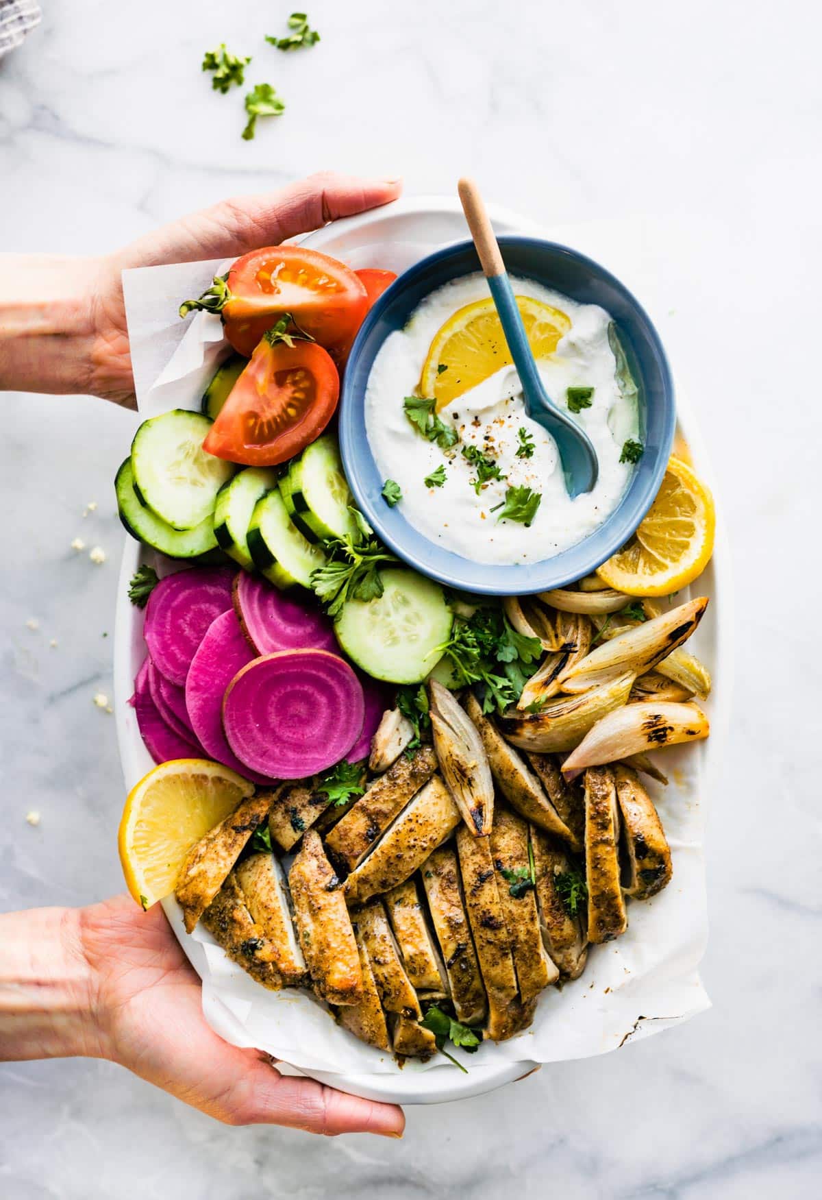 A woman's hands holding a white platter of oven roasted chicken shawarma and shawarma sauce.