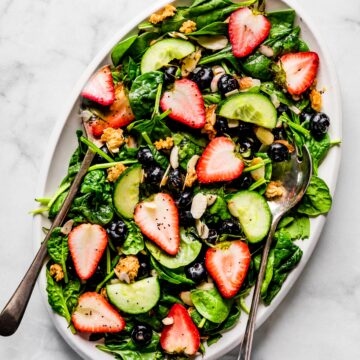 Overhead photo of a strawberry spinach salad on a white platter with salad servers.