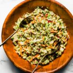 Two metal serving spoons in apple slaw in a wooden bowl.