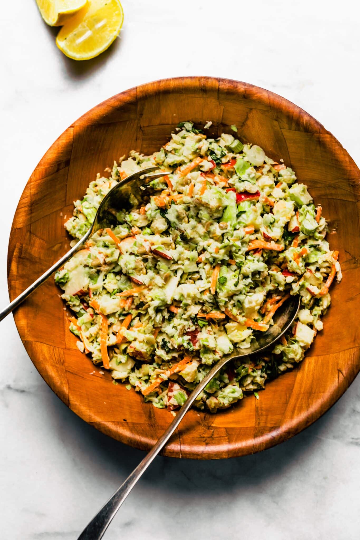 Two metal serving spoons in apple slaw in a wooden bowl.