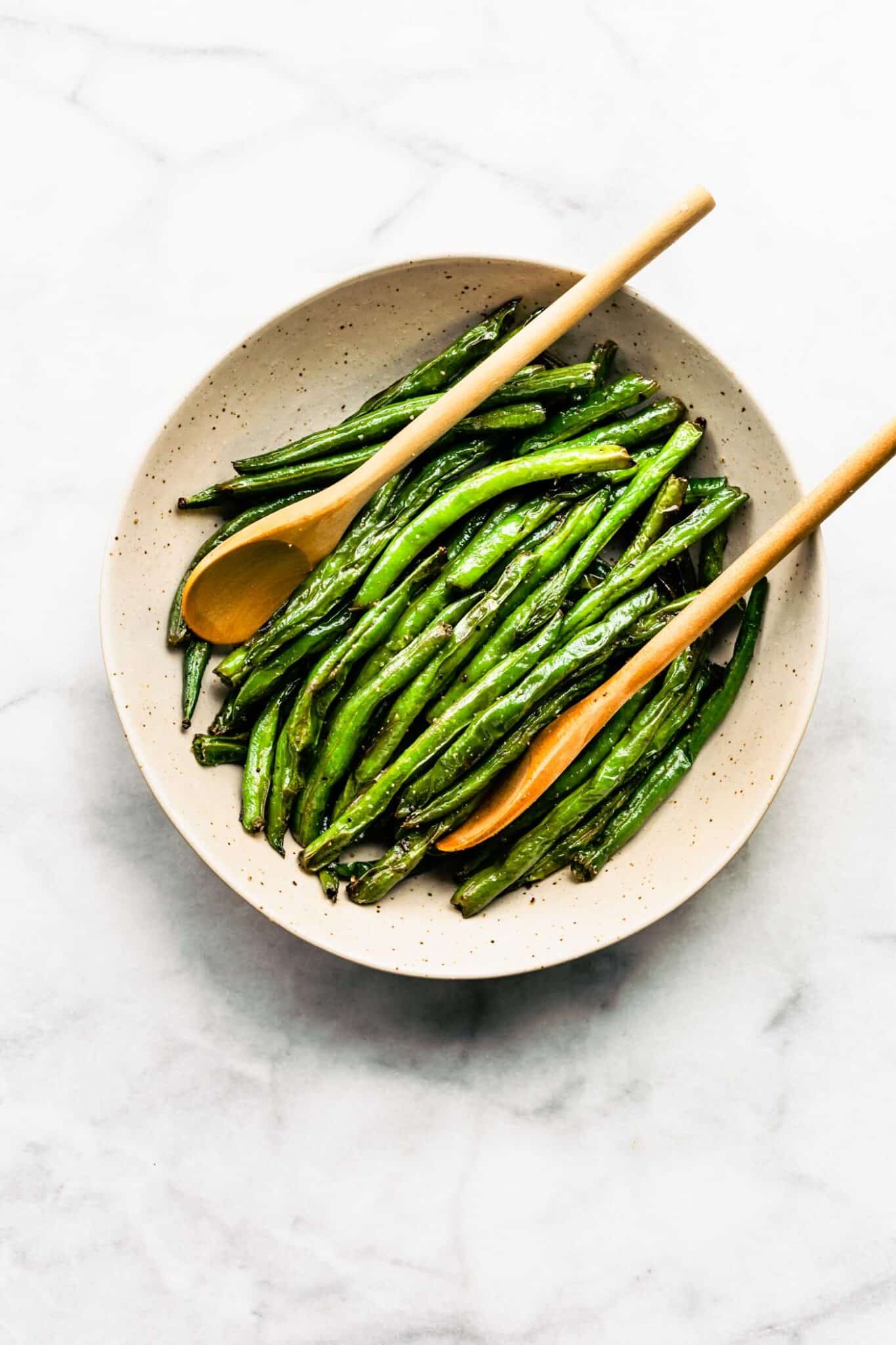 Cooked green beans in shallow bowl with two wooden spoons.