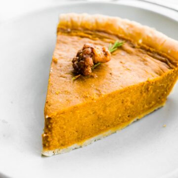 Slice of pumpkin pie on a white plate topped with candied nuts.