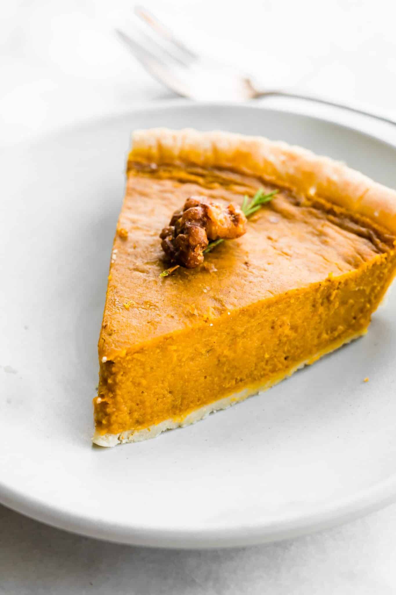 Slice of pumpkin pie on a white plate topped with candied nuts.