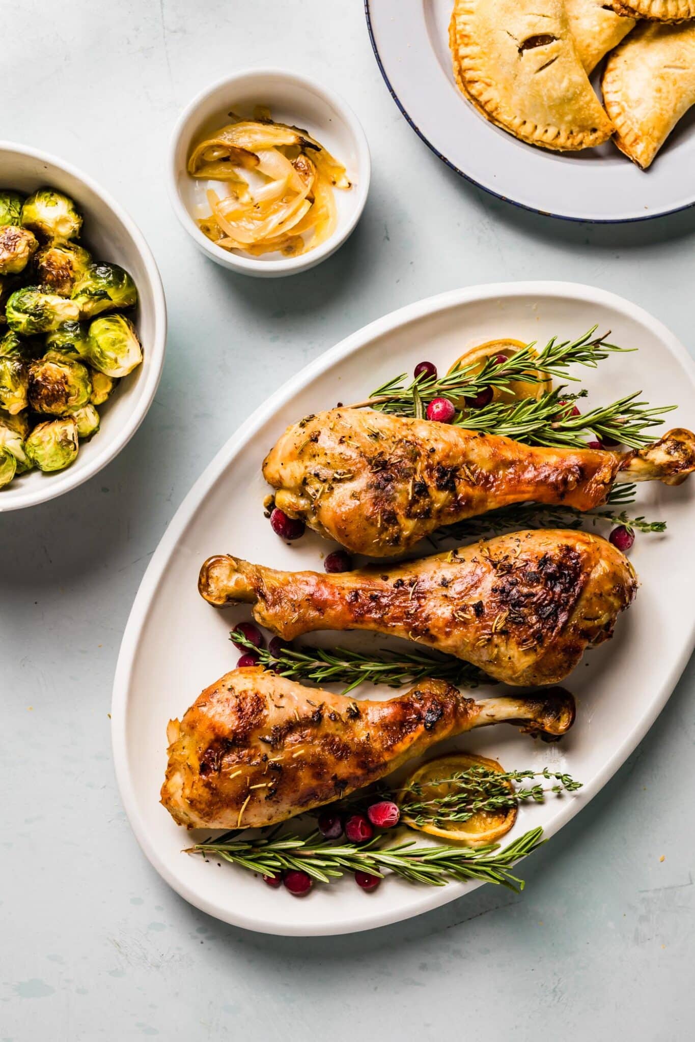 overhead photo of 3 roasted turkey legs on a platter with herbs and lemon slices. 2 other white bowls next to it filled with roasted brussel sprouts, sauteed onion slices, and a plate of gluten free hand pies