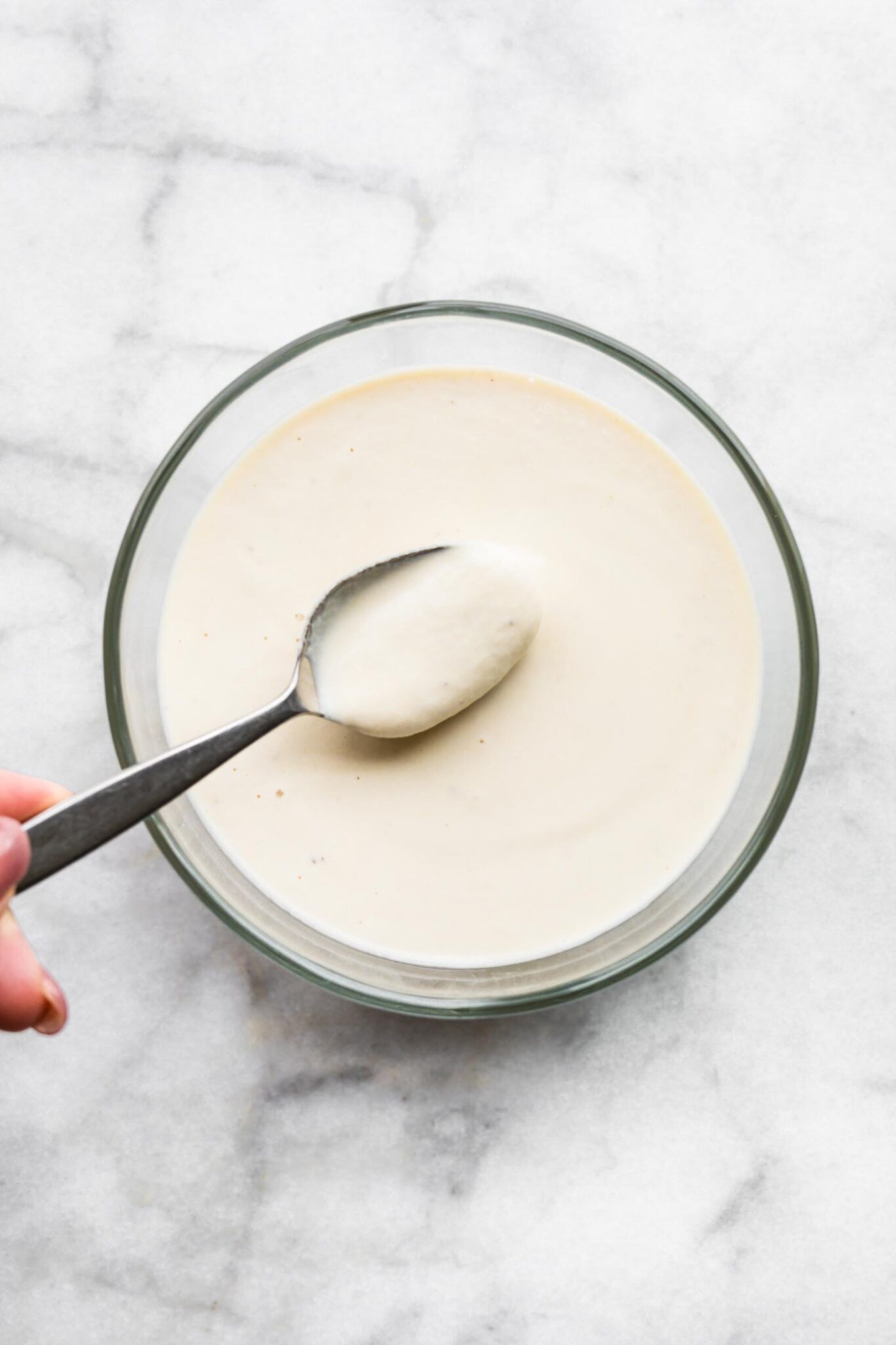 A woman placing spoon in a bowl of cashew cream sauce.