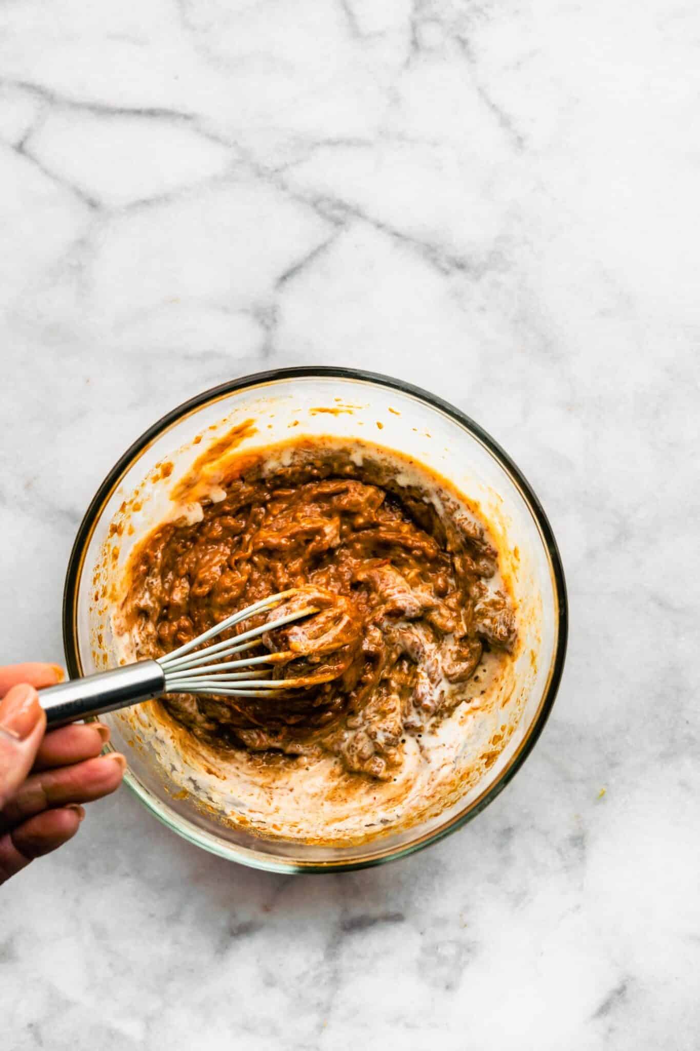 A metal whisk in a glass mixing bowl of peanut sauce ingredients.
