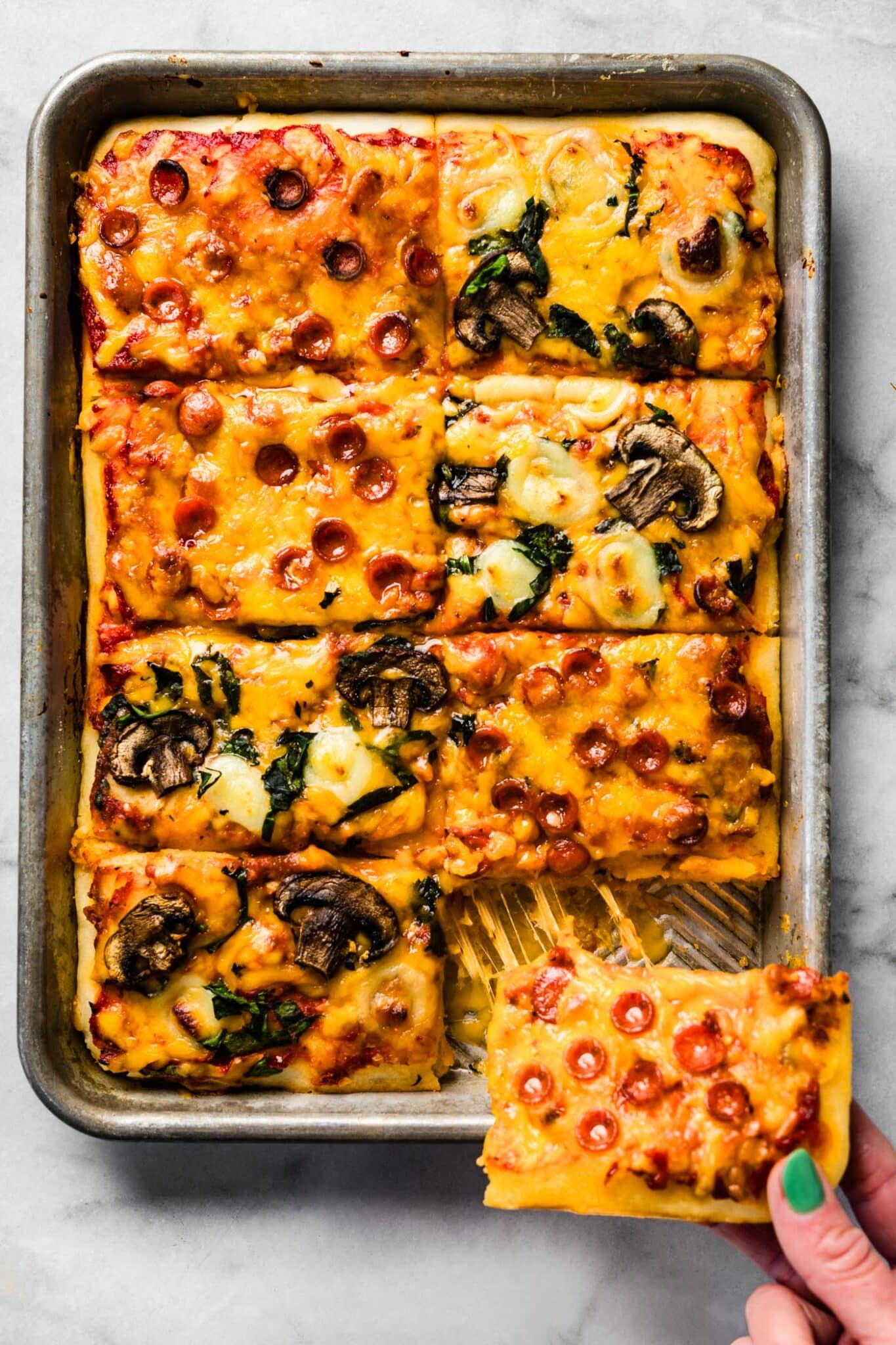 A sheet pan pizza cut into eight slices with a woman pulling a corner piece out.