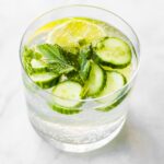 A photo of a short glass of sparkling cucumber water with lemon slice, mint sprig, and cucumber slices garnish