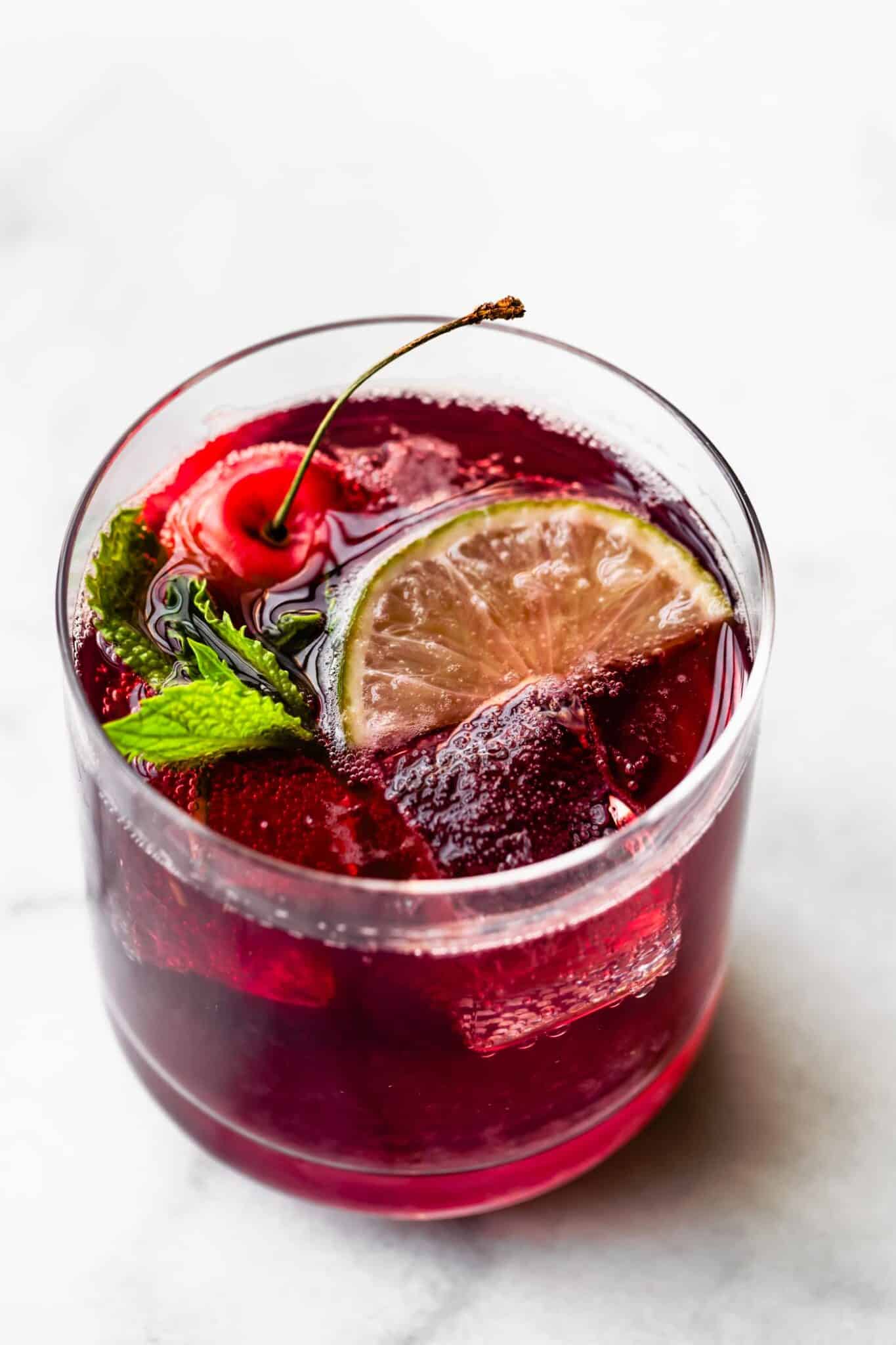 overhead photo of a Clear glass of tart cherry drink garnished with a lime, cherry, and fresh mint.