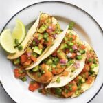 3 shrimp tacos on a white plate topped with avocado and chopped onion and a lime wedge on the plate