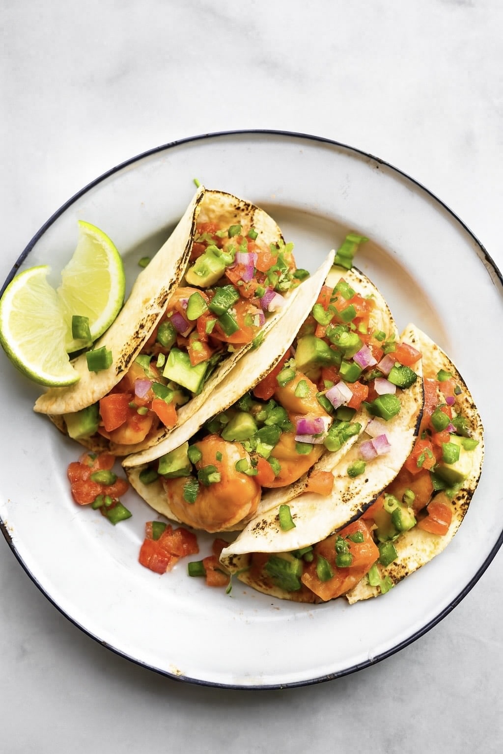 3 fire roasted tomato shrimp tacos on a white plate topped with avocado and pico de gallo served with a lime wedge.