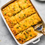 Sliced potato casserole in a white dish with a serving spoon, topped with melted cheese and chopped green onions, with one portion served on a plate.