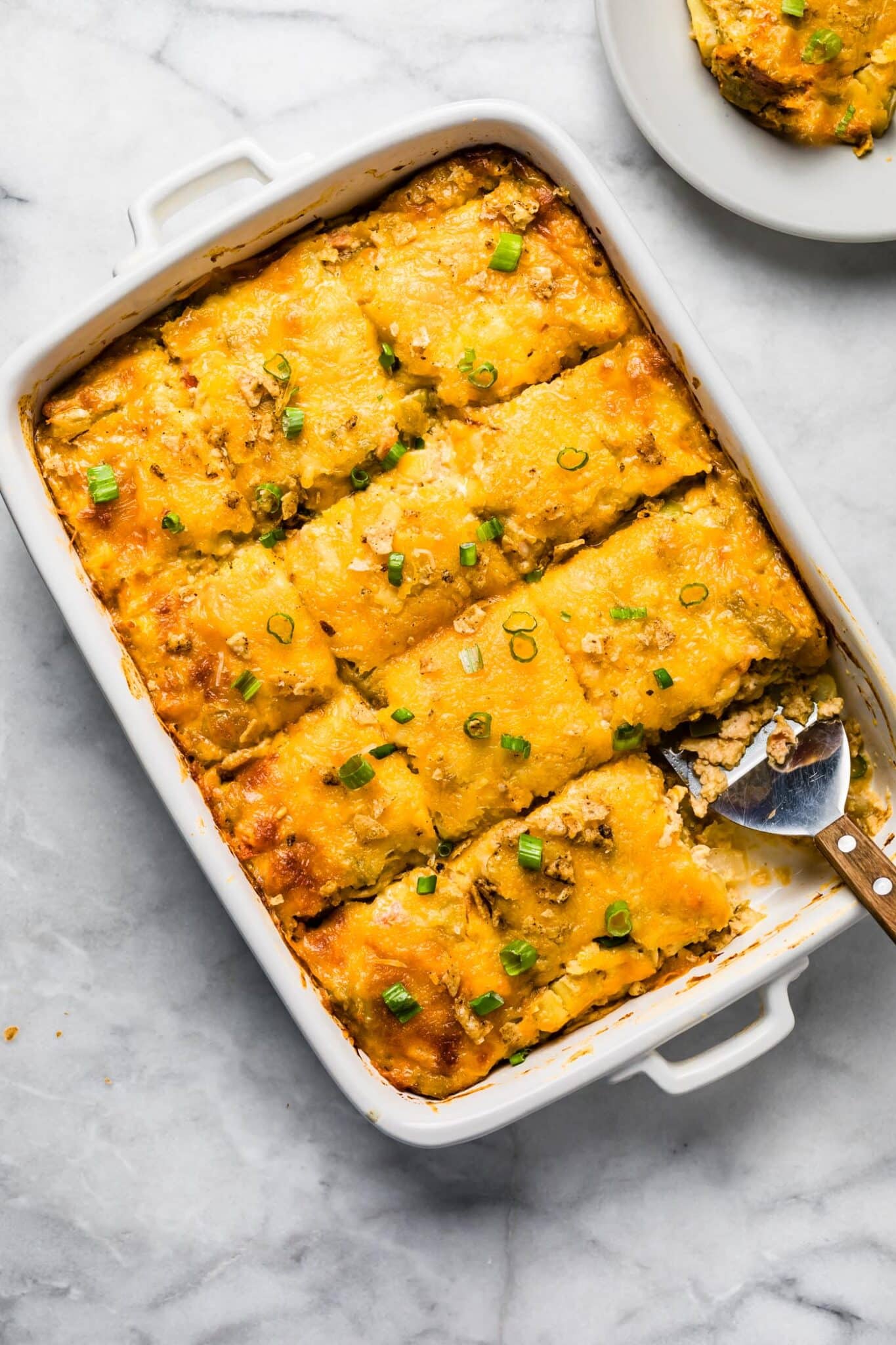 Sliced potato casserole in a white dish with a serving spoon, topped with melted cheese and chopped green onions, with one portion served on a plate.