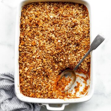Sweet potato casserole in a white baking dish, topped with a golden nutty crumble. A section is scooped out, with the spoon resting in the empty spot. A blue-and-white striped linen sits softly scrunched along the side.