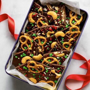 Gift tin lined with parchment paper and filled with Christmas chocolate bark pieces, with a red ribbon beside it.