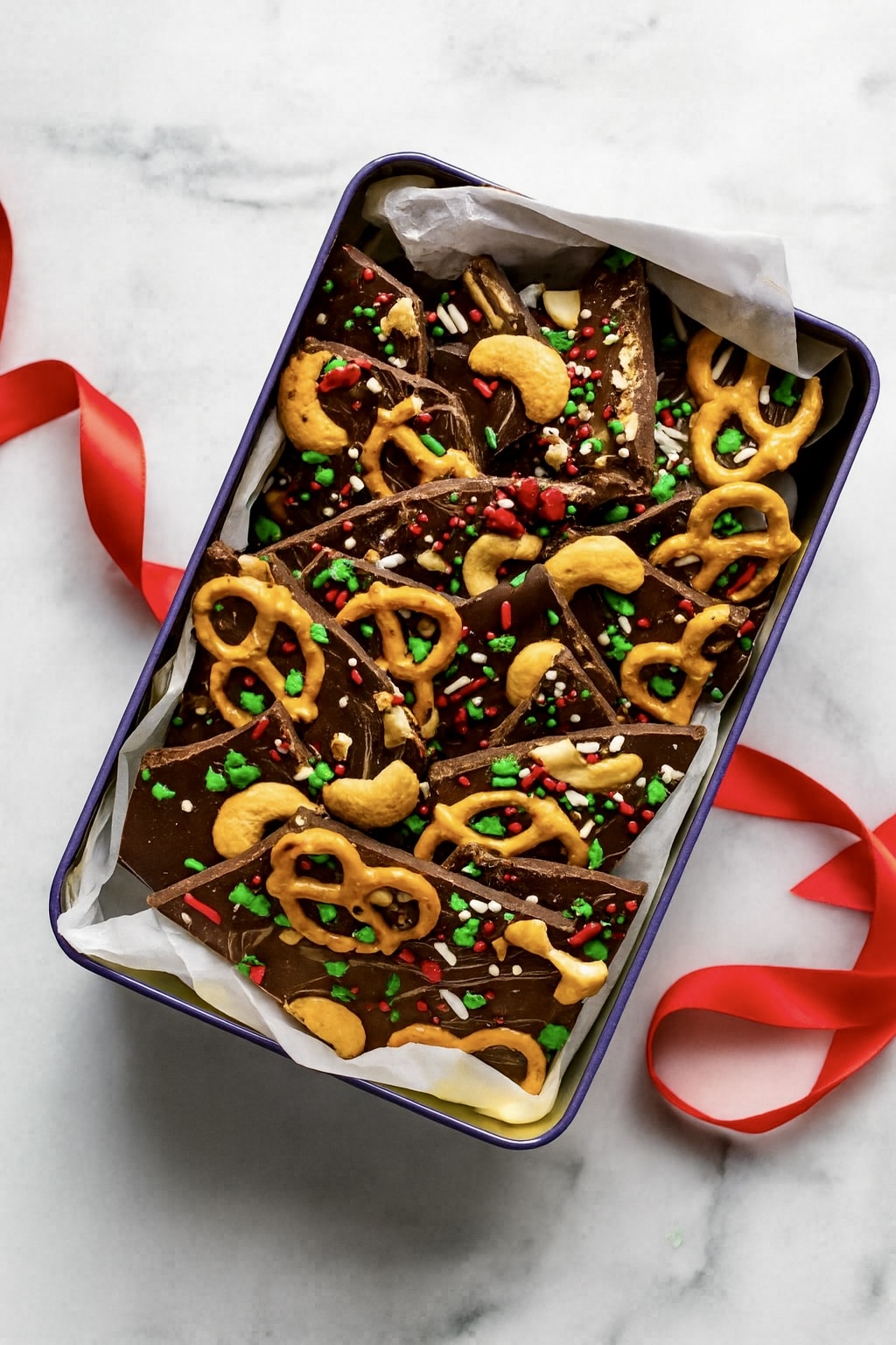 Gift tin lined with parchment paper and filled with Christmas chocolate bark pieces, with a red ribbon beside it.