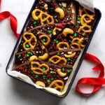 Gift tin lined with parchment paper and filled with Christmas chocolate bark pieces, with a red ribbon beside it.