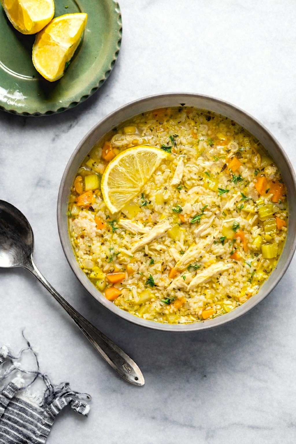 Bowl of lemony chicken and rice soup with vegetables and a lemon slice, with a spoon and extra lemon wedges beside it.