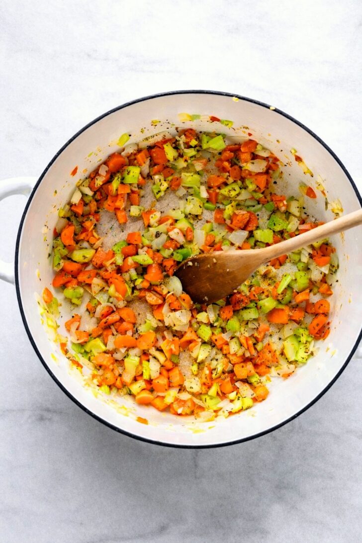 Diced carrots, celery, onions, and garlic sautéing in a white Dutch oven with herbs.