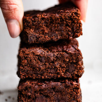 A hand holding a stack of three chocolate black bean brownies, showing a dense fudgy texture and chocolate chips inside