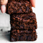 A hand holding a stack of three chocolate black bean brownies, showing a dense fudgy texture and chocolate chips inside