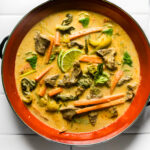 Red pot of finished Thai green curry with beef and vegetables topped with a sliced lime and thai basil