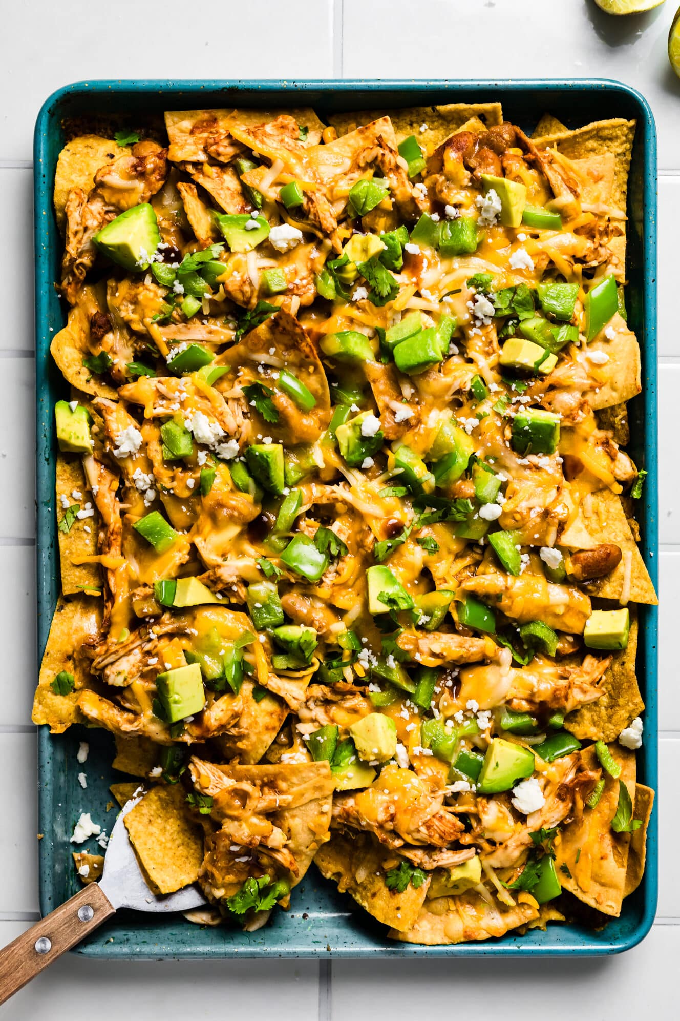 Loaded sheet pan chicken nachos topped with avocado, cilantro, and crumbled cheese on a teal sheet pan with a serving spatula