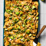 Loaded sheet pan chicken nachos topped with avocado, cilantro, and crumbled cheese on a teal sheet pan with a serving spatula and lime wedges