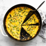 Asparagus frittata sliced in a skillet with a metal spatula and fresh herbs.