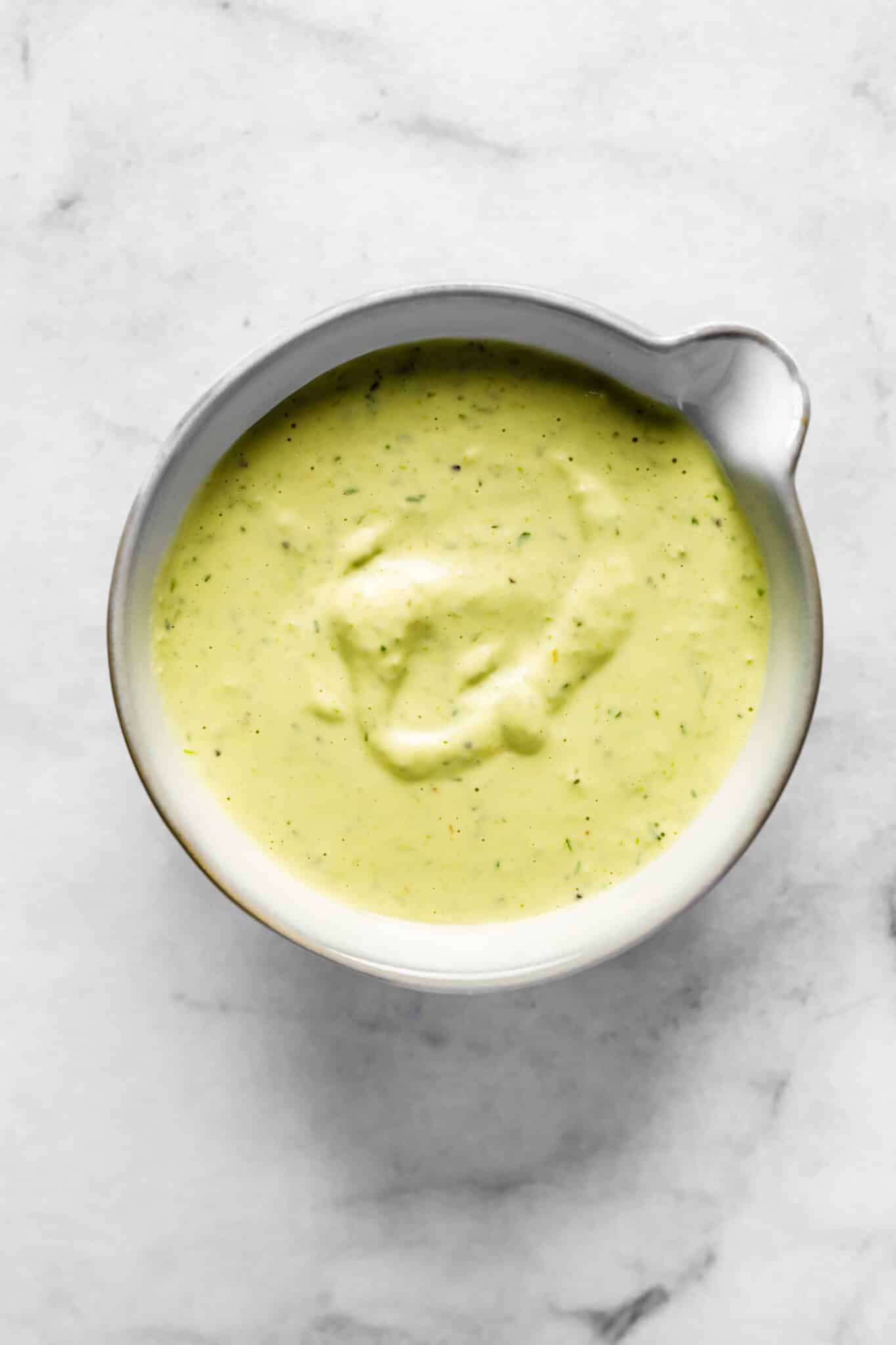 Close-up of creamy pickle dressing in a bowl, with a pale green color and specks of fresh herbs throughout.