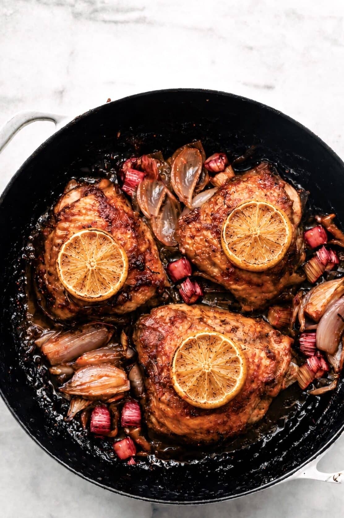 Roasted chicken thighs in a pan with caramelized shallots, chunks of rhubarb, and lemon slices on top.