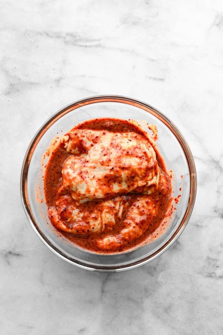 Raw chicken thighs marinating in a red rhubarb BBQ sauce in a glass bowl.