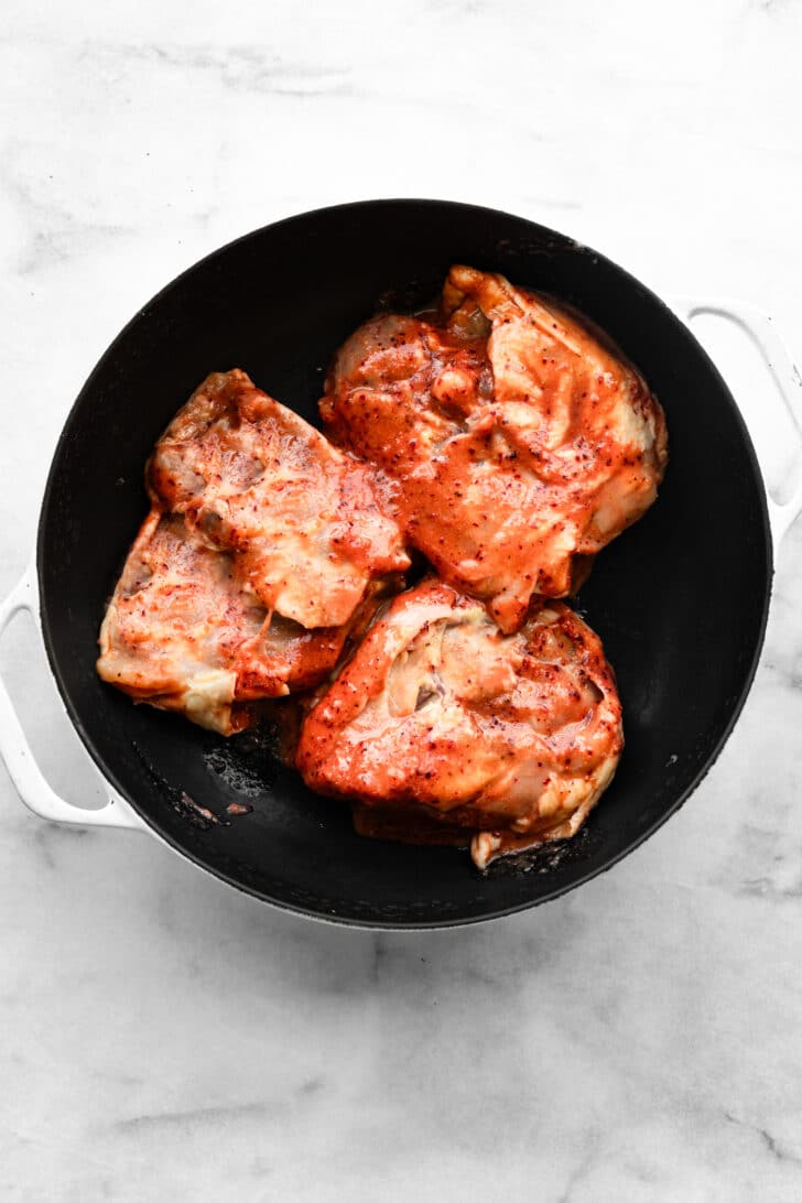 Chicken thighs coated in a red marinade in a skillet (skin side down) before cooking.